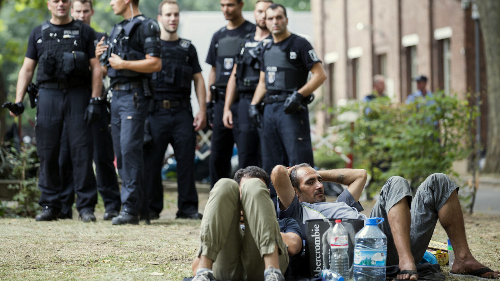 Police stand next to asylum seekers lying on the grass in front of the reception center for refugees in Berlin, Germany, Friday, Aug. 7, 2015. Police was called to support after scuffles between refugees and security officials.